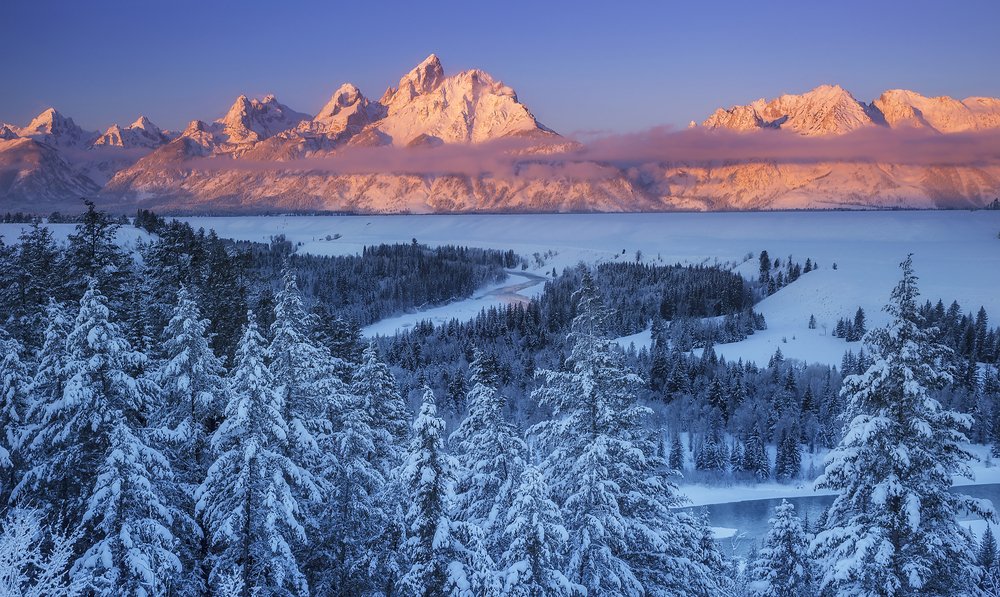 View of the Grand Tetons at sunset with sun colors lighting up the mountains in pastel pink and lavender and a snow-covered landscape everywhere else.