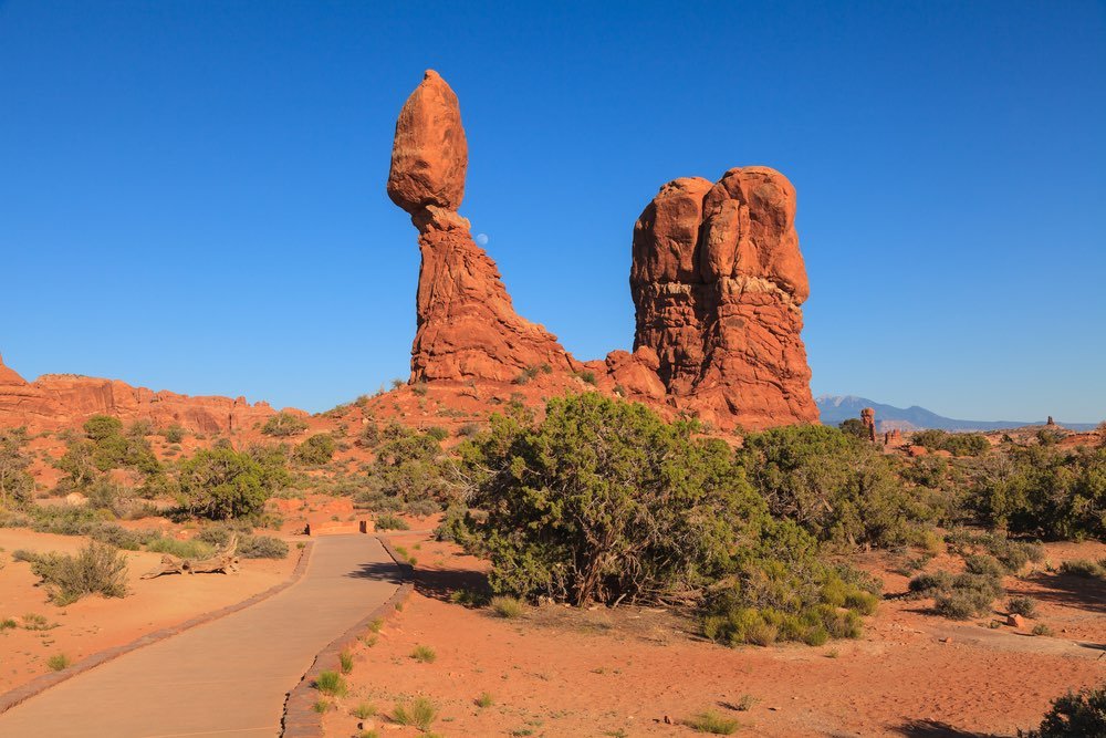 A trail leading up to a red rock formation which features a rock "balancing" on top of another rock, with the moon rising in the background.