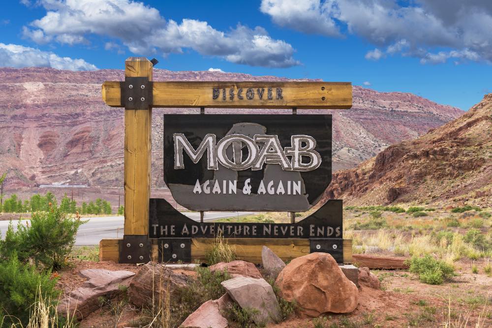 Sign for the town of Moab which reads "Moab Again & Again The Adventure Never Ends" with a desert landscape in a background.