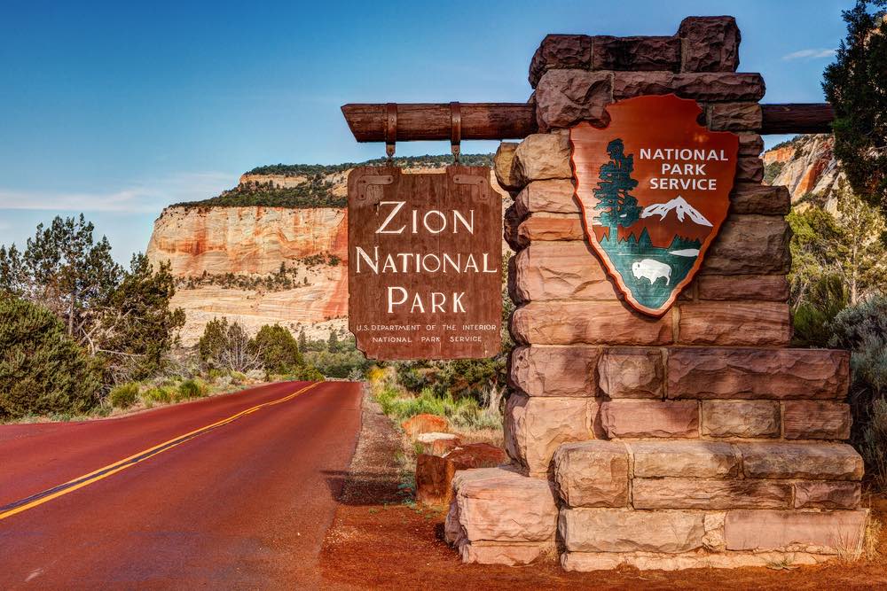 A sign which reads "Zion National Park, National Park Service" on the road leading to the national park with mountains in the background.