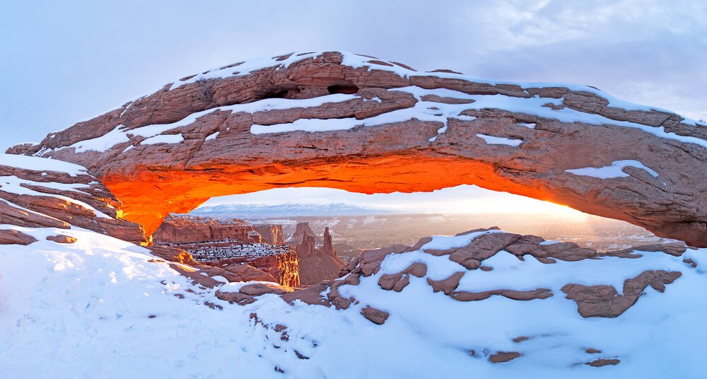 A sunrise view of a snow-covered Mesa Arch illuminating the canyon below, lots of detail covered in snow, on a cloudy day.