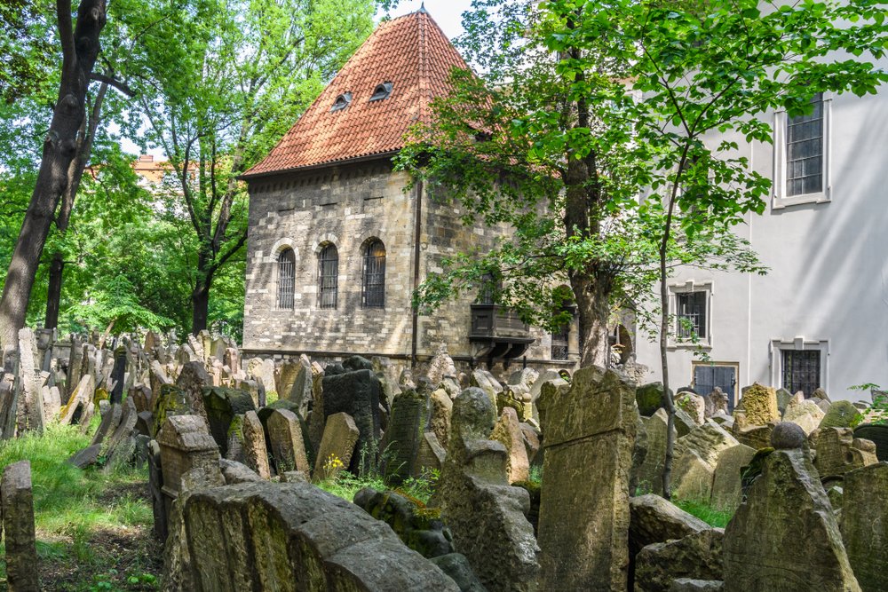 Gravestones stacked up on top of each other in the Old Jewish Cemetery with greenery growing around it, a small building in the middle of the gravestone with a red roof.