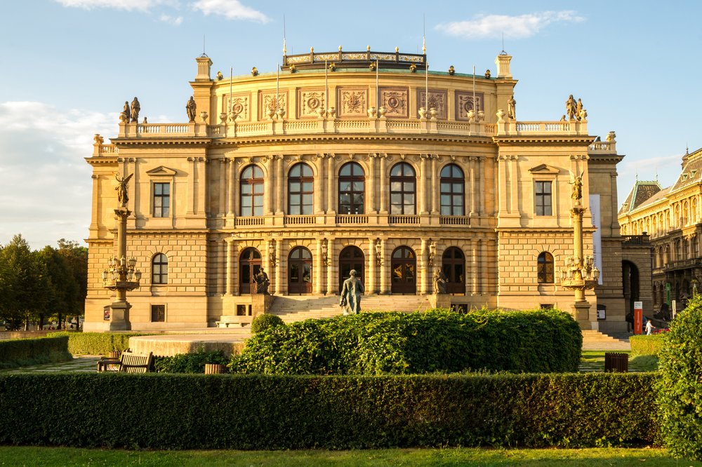 Gold-yellow symmetrical music house with two stories and tons of ornate detailing with a statue in front and green hedges and plants.