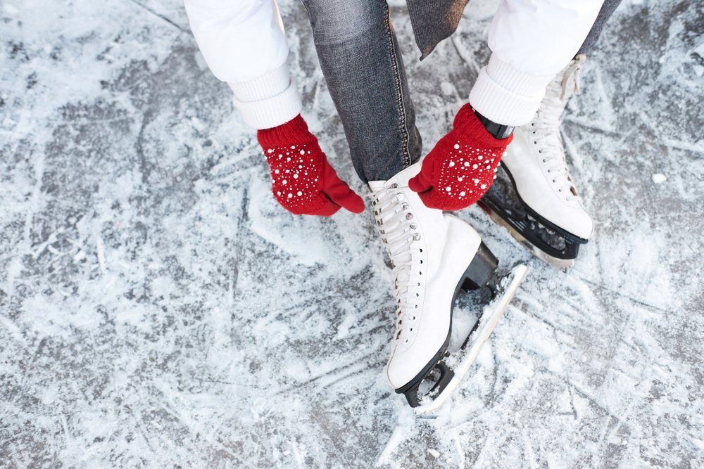 Woman with red gloves, gray pants, white jacket and white skates tying up her laces on the ice.