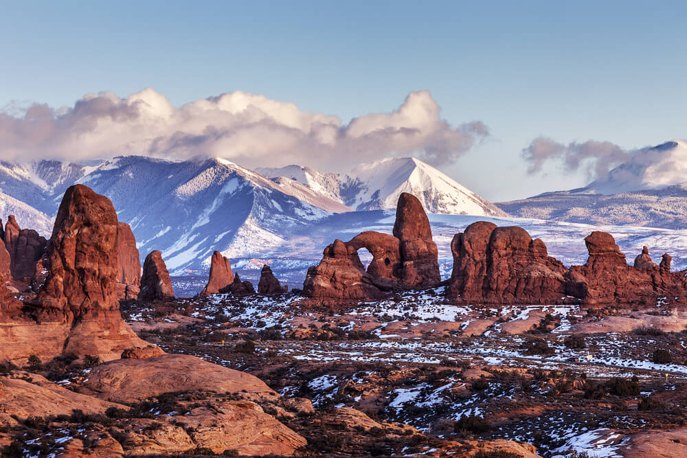 View from afar of the beautiful Turret Arch (which looks like a castle made of natural rock arch) against a backdrop of snow-covered tall mountains, with some light snow on the ground as well.