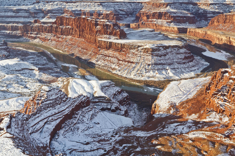 A landscape resembling the Grand Canyon with lots of layered rock carved away by the bend of a river, all the layers of rock are covered in a light snow, alternating orange and white colors.