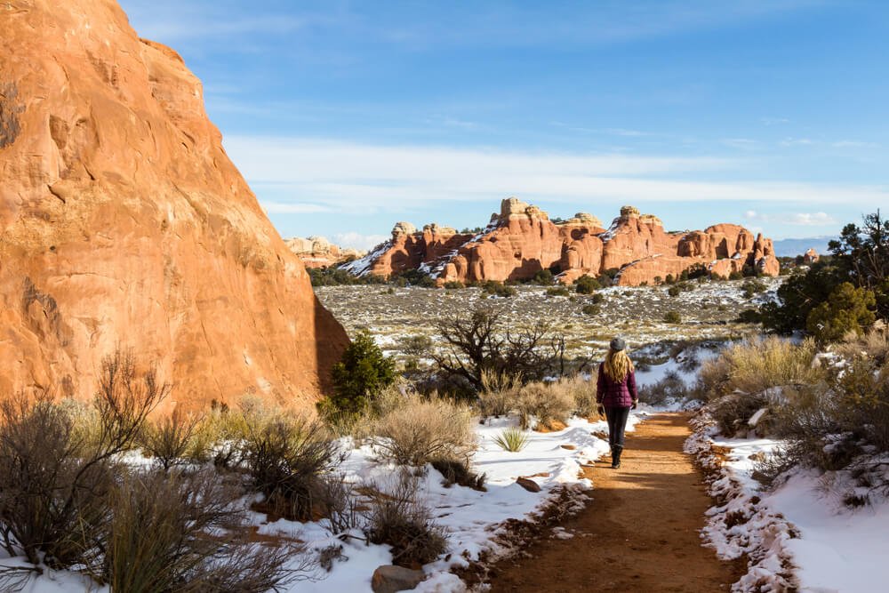 A woman with blonde hair and a maroon jacket walking down a cleared path with snow on each side in Arches National Park winter landscape of snow and red rocks.