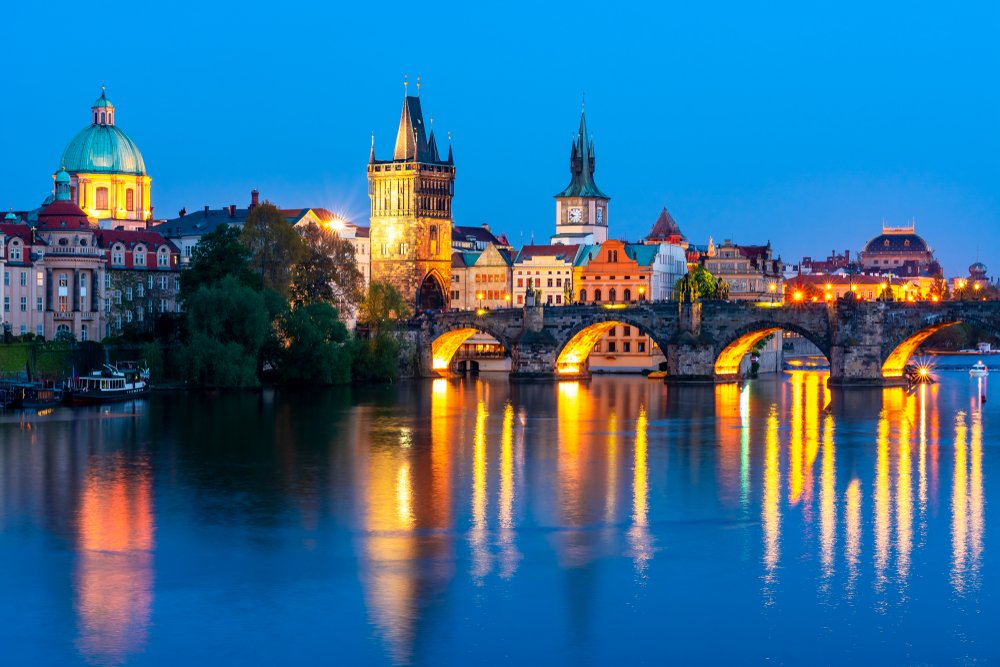 Blue hour in Prague with lots of lights on in the city, reflecting in the water, you can see Charles Bridge, a watchtower, and distinctive Prague architecture in the skyline.