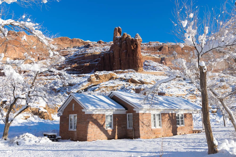 A small red brick building covered in snow surrounded by a red rock landscape with a light dusting of snowfall.