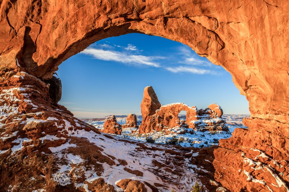 Snowy view of Arches National Park in the winter, looking through one arch and seeing a larger complex of arches through the "window", red rock covered in snow.