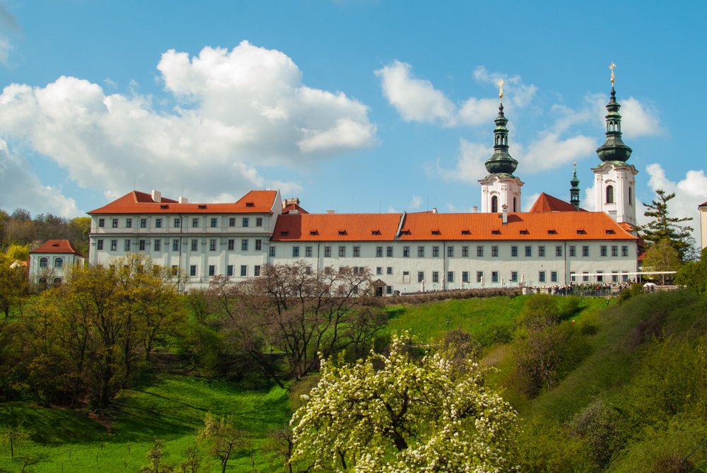 A view from the side of Strahov monastery, a white monastery with a red tiled roof and two towers, on the top of a small hill.