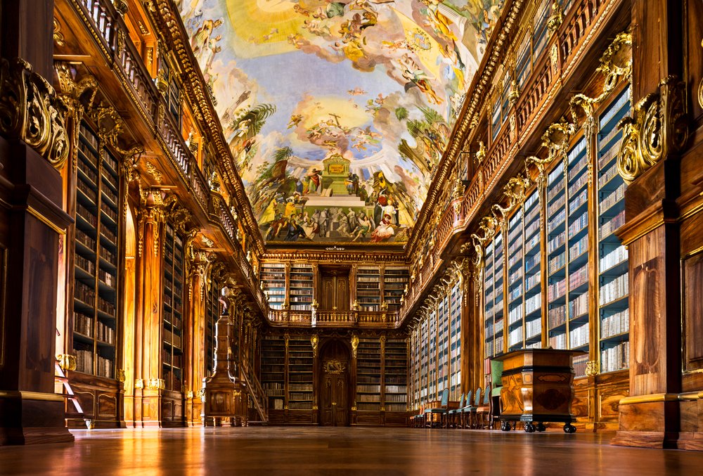 An ornate library room with a pastel painted ceiling with lots of detailing and rows upon rows of books with wooden shelves and carved wood detail.