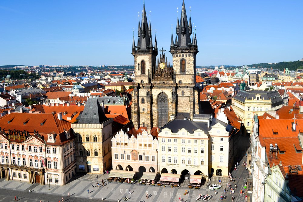 The view of the famous Prague church from a high vantange point, people down in the square looking very small, with lots of red roof architecture and pastel building facades surrounding the church in the middle of the photo.