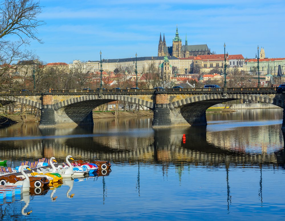 View of Legion Bridge from the water with a view of some novelty water boats (such as swan boats) in the water. Prague Castle is in the background.