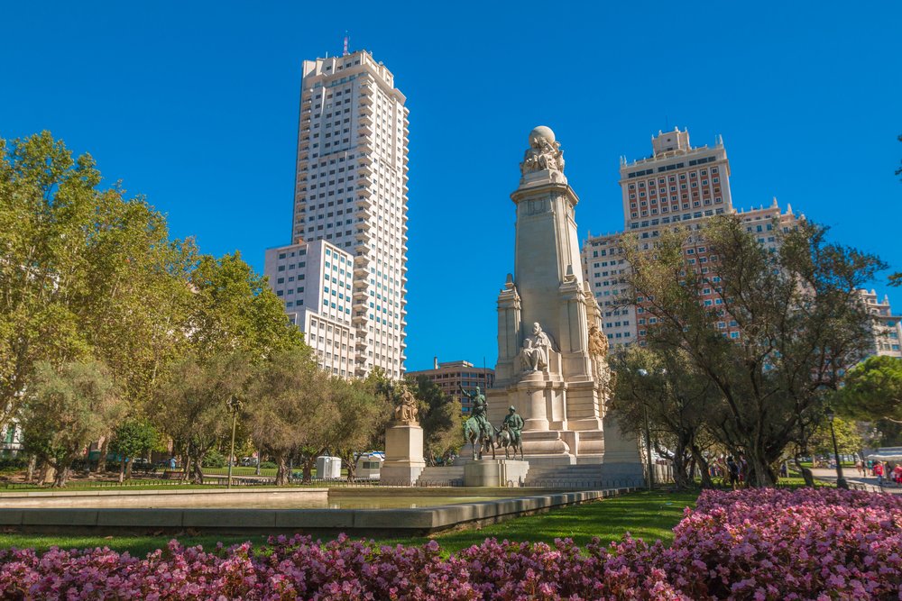 A giant statue in the middle of the park with two skyscrapers around it.