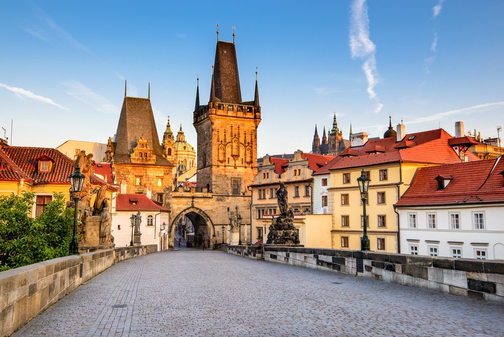 Walking across the Charles Bridge towards Mala Strana and the Castle District, buildings with red roofs on the side and observation towers and a basilica dome in the background.