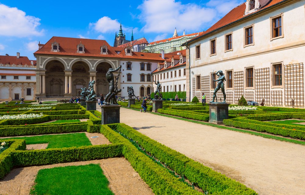 Interior landscaped gardens of the Prague Castle, with statues, green low hedges, buildings with interesting tile work, and red roofed architecture.