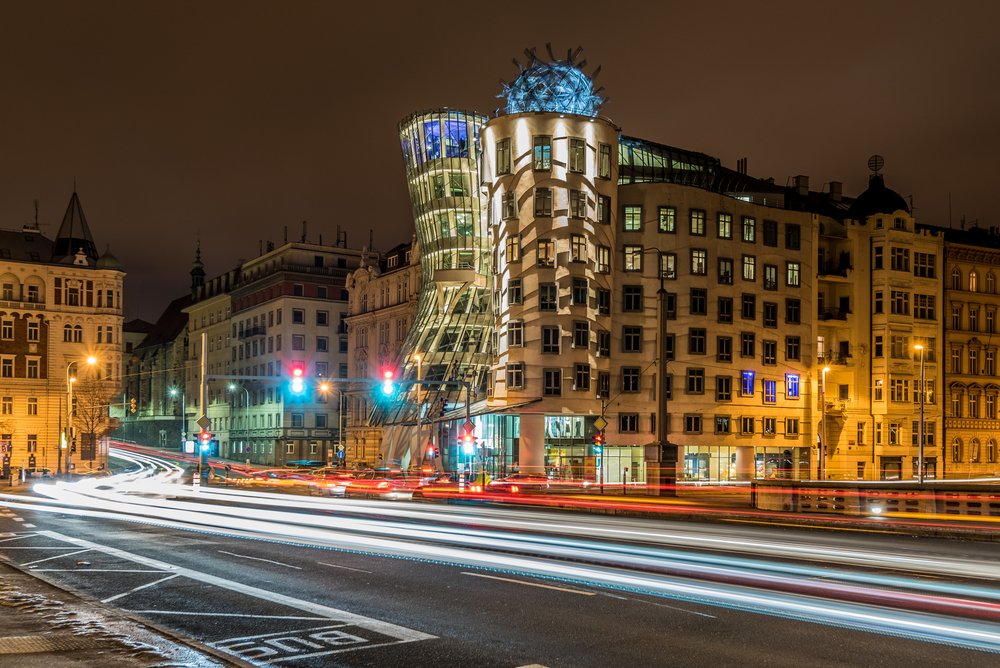 The Dancing House building lit up at night: two buildings intertwined together with  a movement that makes them look like they're dancing, with light trails on the street in the foreground.