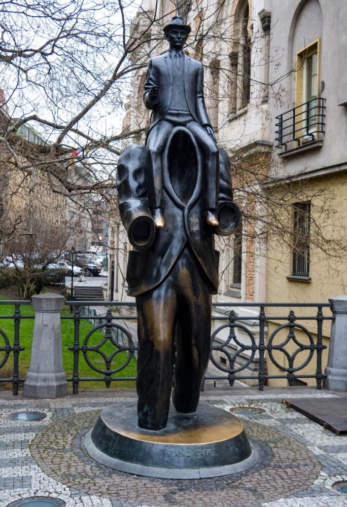 Bronze statue of a headless, armless man with a man with a hat sitting on top of his shoulders, with a city background behind the sculpture.