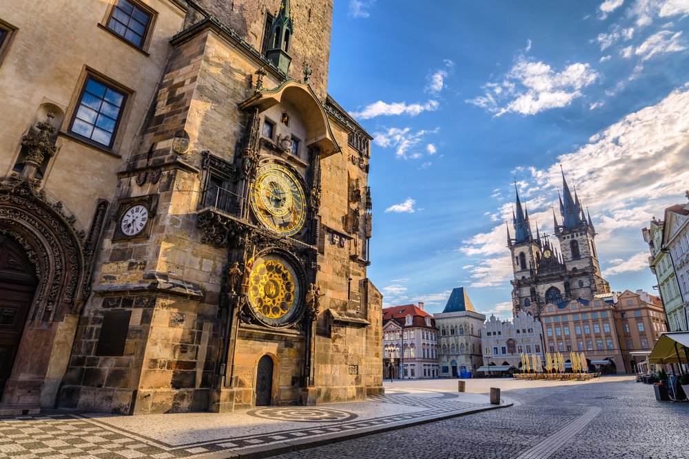 The Old Astronomical Clock with lots of painted detail and on the right side the Our Lady Before Tyn Church with two distinct spires, lots of pastel buildings around the square with cobblestone walkways.