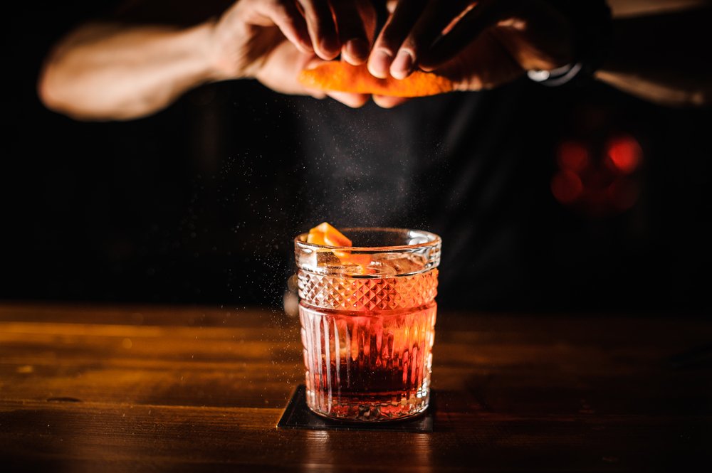 A man's hands making a cocktail, which looks to be a whiskey old fashioned, with an orange peel garnish, in a dark bar.