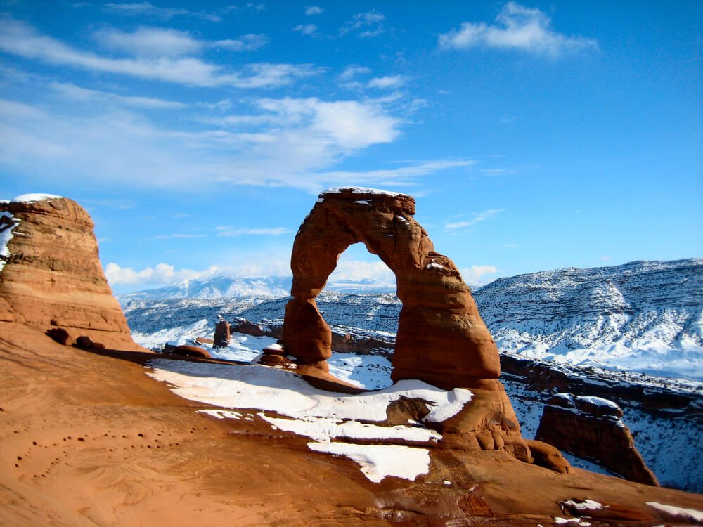 A view of a single orange sandstone arch against a backdrop of snow-covered mountains and a blue sky.