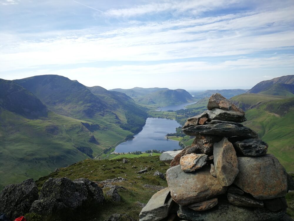 A rock cairn overlooking green fells and small hills and a blue lake in the Lakes District