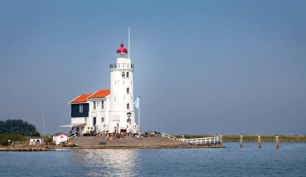 A white lighthouse with an orange roof and black room at the back of the lighthouse, shown on the water, a true hidden gem in Amsterdam.