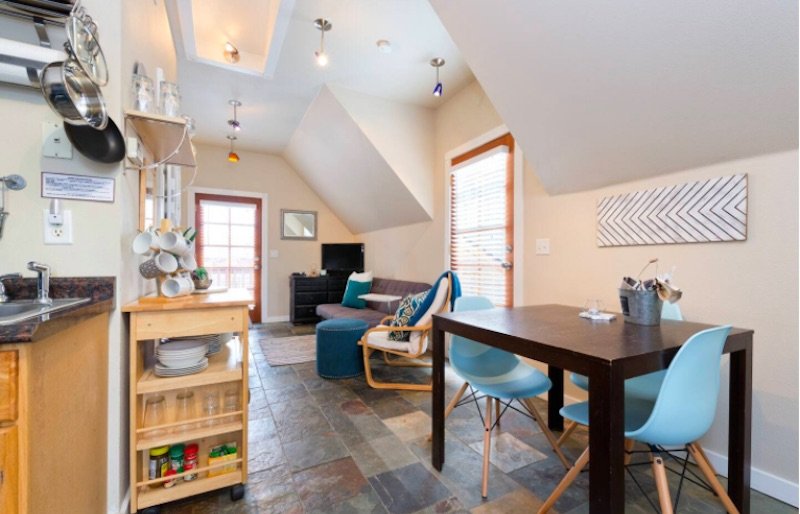 View of a spacious modern Airbnb loft with kitchen equipment, table for two with blue chairs, and a couch seating area with white walls and modern design.