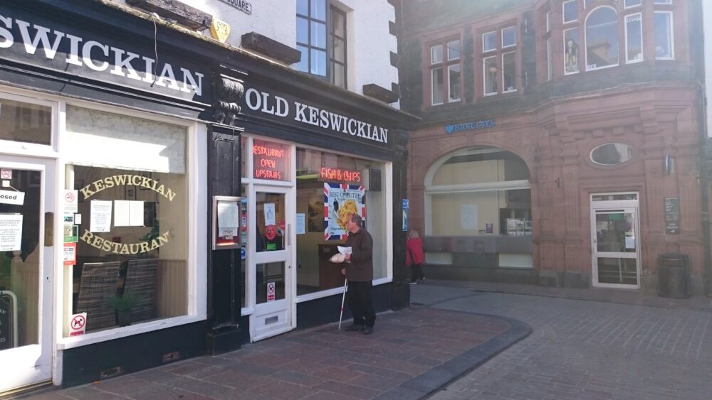 A traditional high street in England with a man looking inside the restaurant that says "Old Keswickian" and has posters for fish and chips.