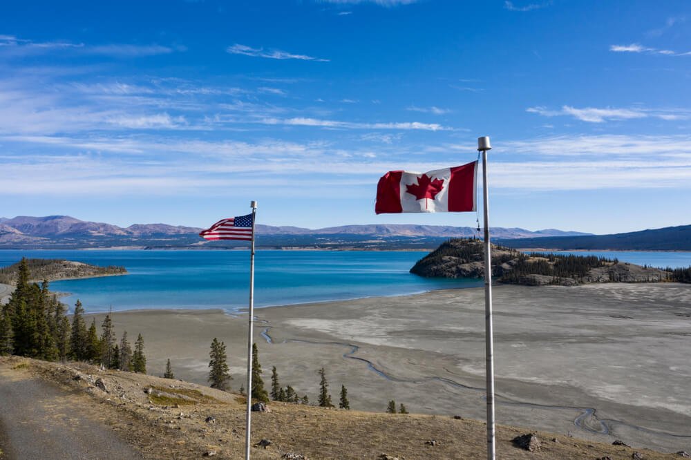 An American and a Canadian flag side by side next to barren fields and a deep blue lake while driving the Alcan Highway road trip to Alaska