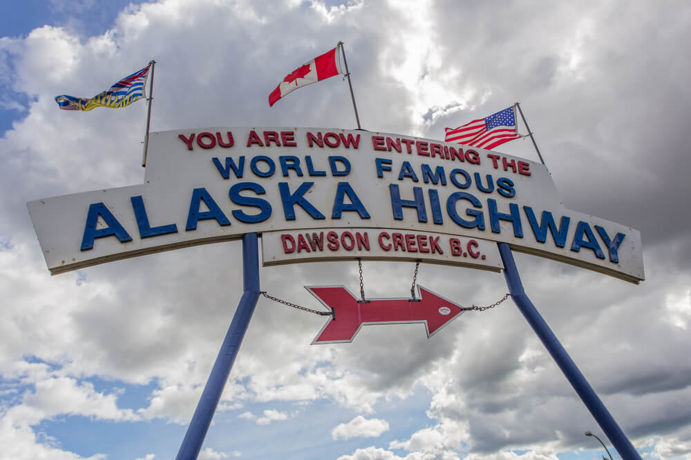 Sign with three flags reading "You are now entering the World Famous Alaska Highway Dawson Creek BC" 