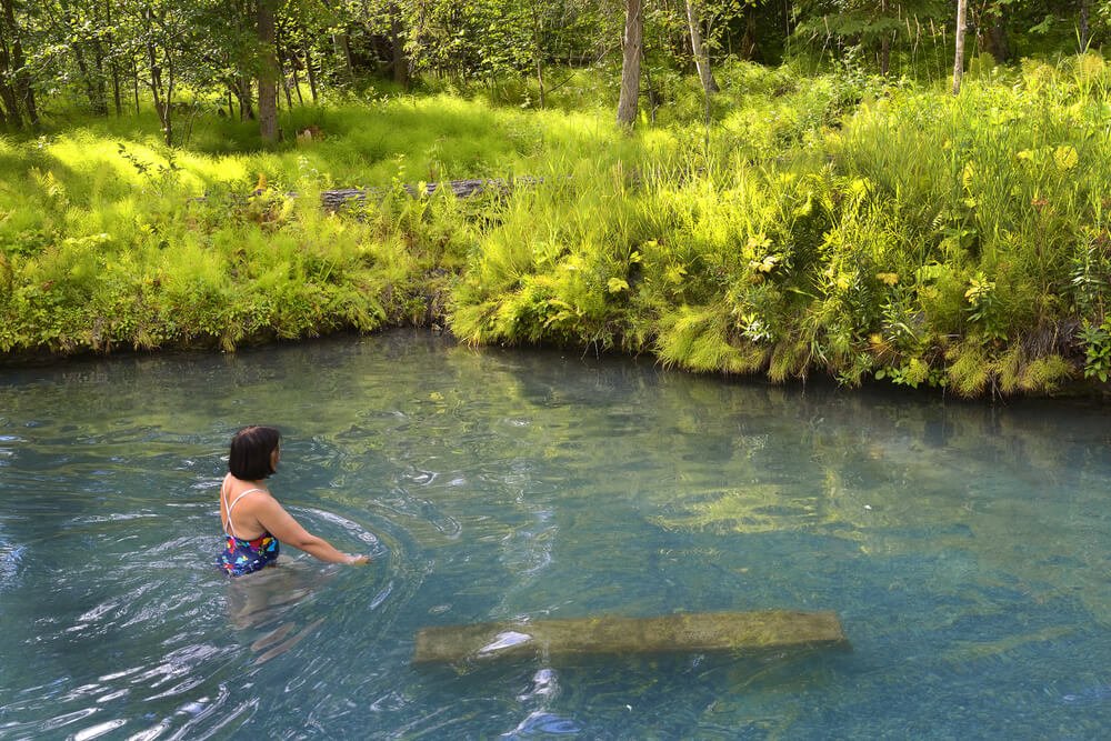 Woman with short brown hair in a colorful bathing suit in turquoise water at a hot spring in Canada surrounded by green grass and trees.