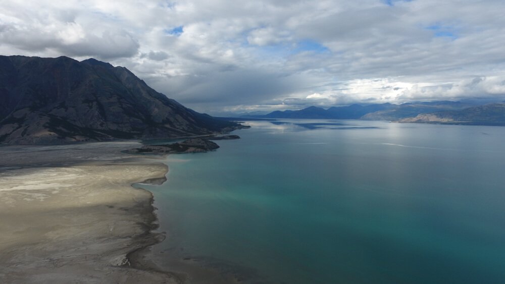 Soft blue water against beige sand in a bay in Canada, with larger hills in the distance and lots of cloud.