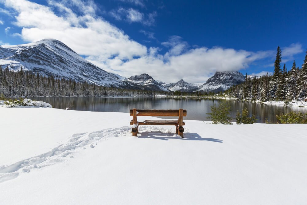 An empty bench with footstep tracks next to it, looking out over Lake McDonald and all the snow-covered mountains surrounding it.