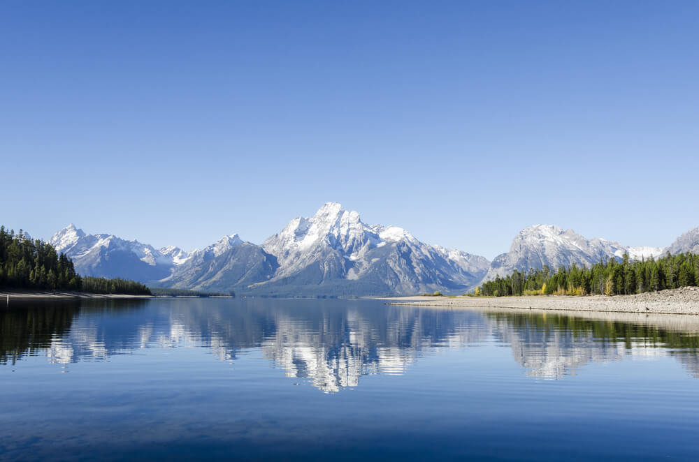 A mostly still lake reflecting the mountains of the Teton Range beautifully in its glassy, slightly rippled surface.