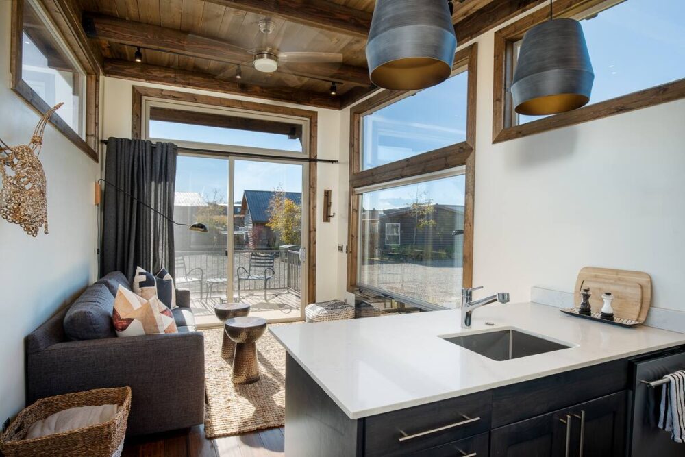 A tiny house with a small kitchen, small coach, and large windows overlooking a nature landscape.