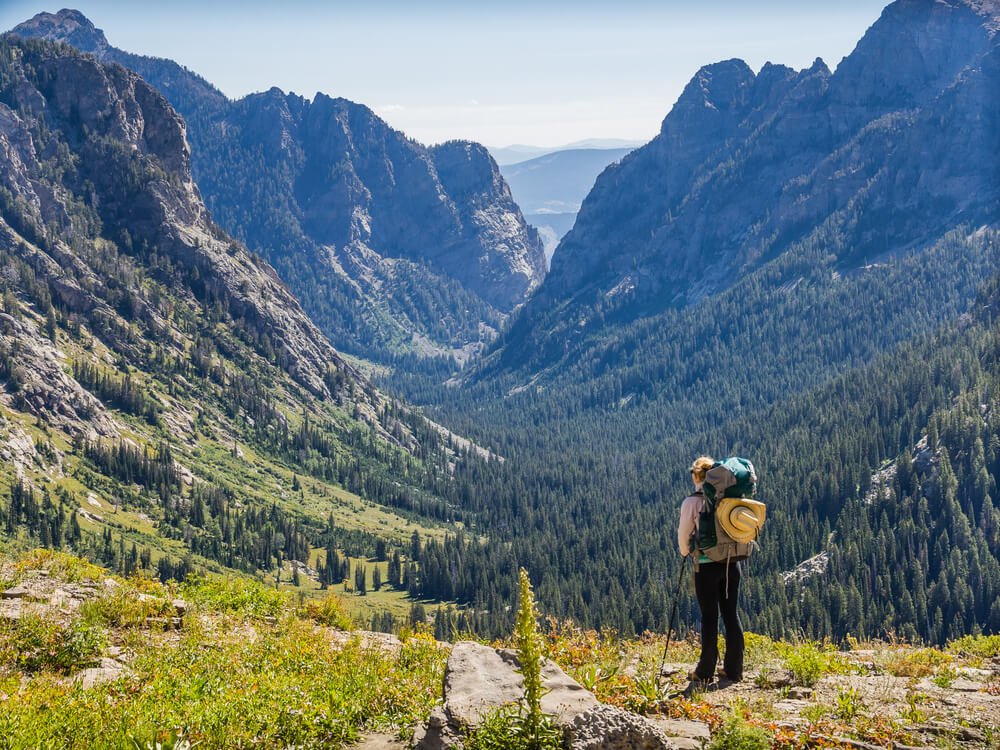 A female hiker looking at a valley in Grand Teton National Park, well-prepared with a backpack, hiking poles, and a sunhat on her back.