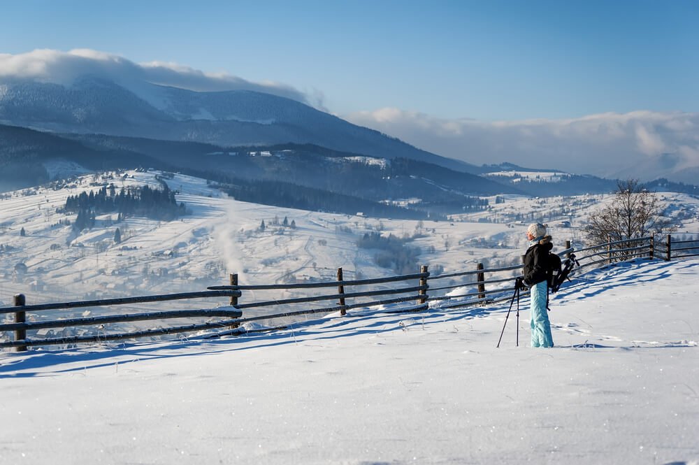 Female hiker in snowshoes with trekking poles wearing a black jacket and baby blue pants, looking over a snow-covered mountainous landscape in Glacier National park in the winter time.