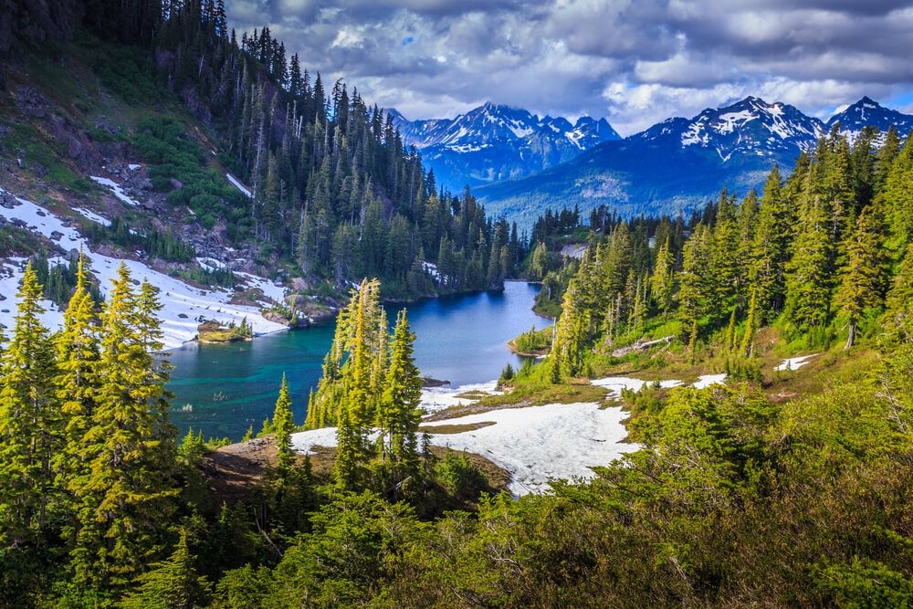 An overcast day in Glacier National Park, turquoise water next to melting snow, surrounded by evergreen trees and mountains with little snow.