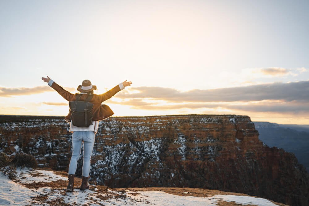 Woman wearing hat, jacket, jeans, and snow boots at the Grand Canyon in winter, with her arms up in the air as the sun sets.