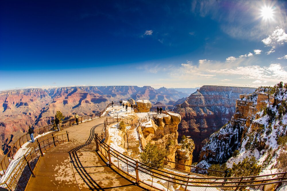 Icy trails at Grand Canyon in winter, with snow on the sides and in the canyon itself interspersed with red rock, with visitors at the end of the trail.