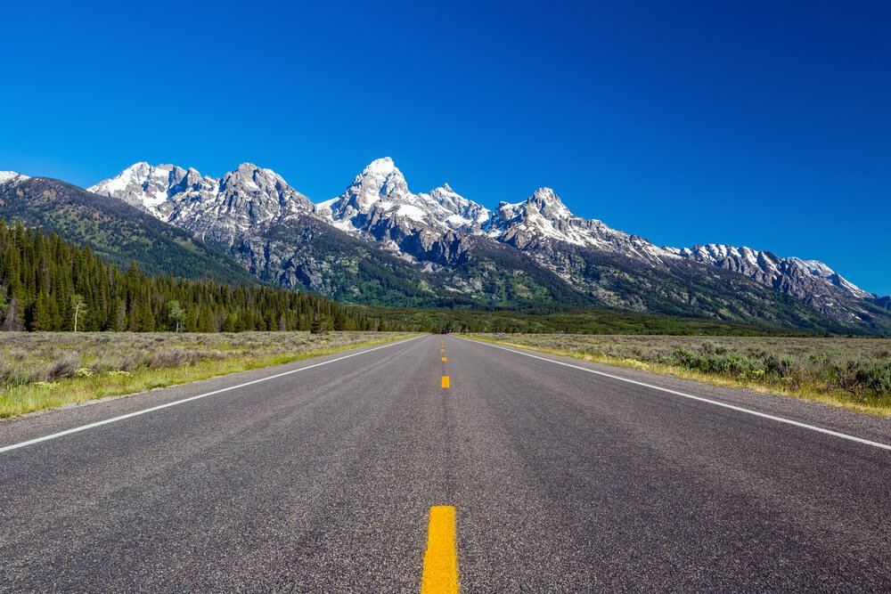 An empty road leading towards the distinctive peaks of the Teton Range near Grand Teton National Park on a cloudless summer day.