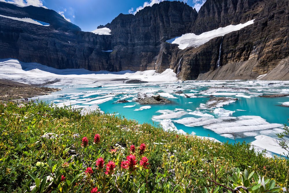 Brilliant turquoise blue water surrounded by white glacial ice, with green grass with red wildflowers on the edge, surrounded by tall mountain edges lightly covered in snow.