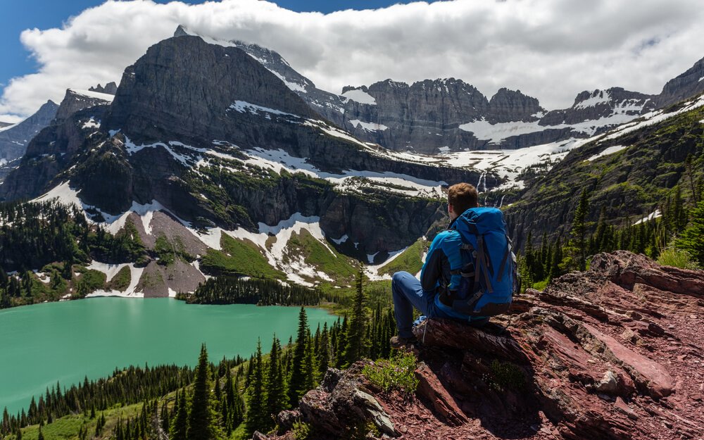 Male hiker wearing blue jacket and blue backpack sitting after doing some Glacier National Park hiking, looking over the teal colored Grinnell Lake, surrounded by mountains dusted with some remaining snow.