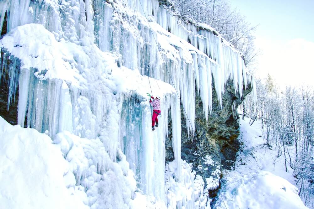 A woman in a pink jacket and red pants climbing up the icicles of a frozen waterfall with climbing equipment and harness.