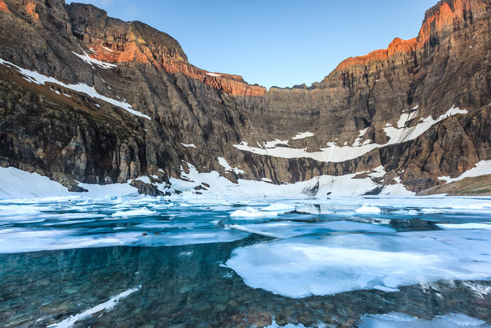 Blue glacial water topped with some unmelted glacial ice, surrounded by reddish-brown rocks lit up orange-red by the last of the afternoon light, on a popular hike in Glacier National Park