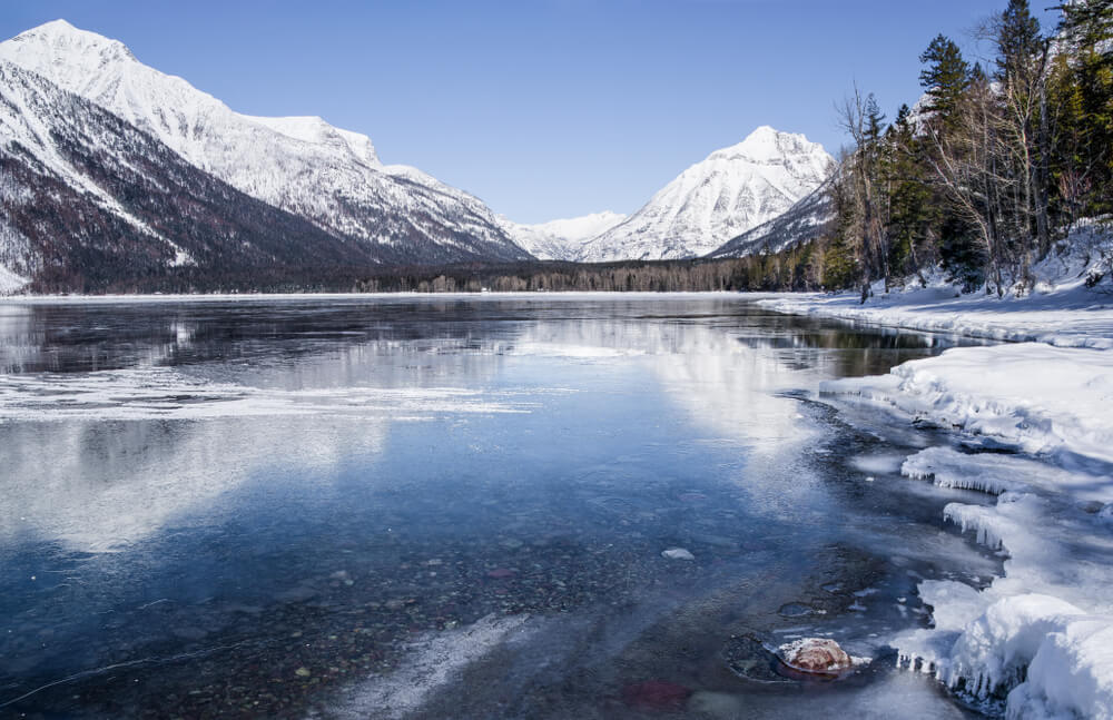 Icy surface of Lake McDonald as seen in winter, wth snow and ice on the banks of the lake, with snow-covered mountains in the distance.