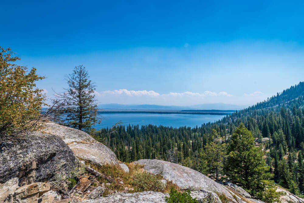 Giant boulders in front of the view from Inspiration Point, showing lots of evergreen trees in front of a brilliant blue lake with some clouds on the horizon but an otherwise clear sky.