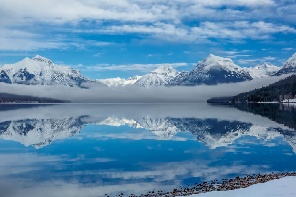 The unfrozen surface of Lake McDonald in early winter in Glacier National Park, reflecting the snow-covered mountains with a patch of fog on a sunny winter day.